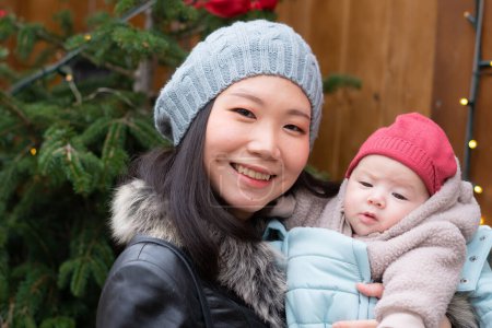 Téléchargez les photos : Jeune femme coréenne asiatique heureuse et attrayante tenant bébé fille dans ses bras devant l'arbre de Noël marchant sur le marché de rue d'hiver dans le concept d'amour mère et fille - en image libre de droit