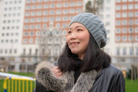 Téléchargez les photos : Portrait urbain en plein air de jeune femme chinoise asiatique heureuse et belle en manteau et bonnet en cuir faisant une promenade détendue dans le quartier des affaires de la ville souriant gai - en image libre de droit