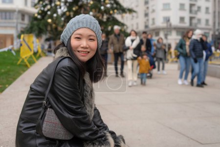 Téléchargez les photos : Portrait urbain extérieur de jeune femme coréenne asiatique heureuse et belle en manteau et bonnet en cuir faisant une promenade détendue dans le quartier des affaires de la ville souriant gai - en image libre de droit