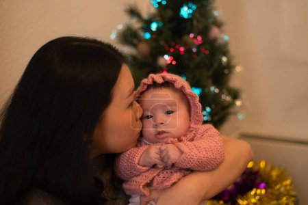 Téléchargez les photos : Lifestyle portrait of young attractive and happy Asian woman in front of Christmas tree  holding baby girl in her arms, her adorable and sweet little daughter - en image libre de droit