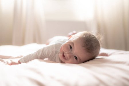 Foto de Adorable y feliz niña de 5 meses descubre alegremente alegría ilimitada mientras explora juguetonamente su cama. Rodeado por el cálido resplandor de la luz natural, el pequeño sonríe alegremente - Imagen libre de derechos