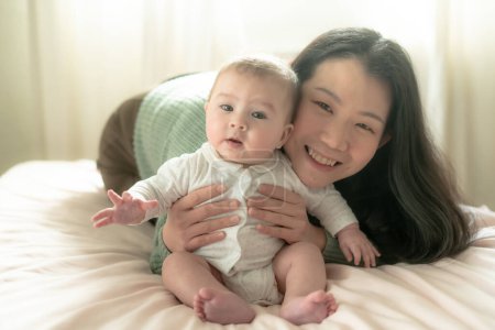 Foto de Amor y alegría, una joven coreana asiática se involucra alegremente con su bebé en la cama. Su risa crea recuerdos preciosos unidos, unidos en pura felicidad. - Imagen libre de derechos