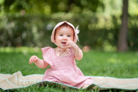 Foto de Adorable y feliz niña al aire libre en el parque - retrato de 7 u 8 meses de edad hermoso niño sonriendo alegre sentado en la estera en la hierba en el parque de la ciudad con sombrero acogedor y vestido - Imagen libre de derechos