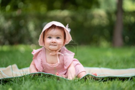 Foto de Adorable y feliz niña al aire libre en el parque - retrato de 7 u 8 meses de edad hermoso niño sonriendo alegre sentado en la estera en la hierba en el parque de la ciudad con sombrero acogedor y vestido - Imagen libre de derechos