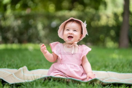 Photo for Adorable and happy baby girl outdoors in the park - portrait of 7 or 8 months old beautiful little child smiling cheerful sitting on mat on grass at city park wearing cozy hat and dress - Royalty Free Image