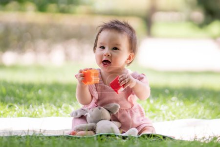 Foto de Adorable y feliz niña jugando al aire libre en el parque - retrato de 7 u 8 meses de edad hermoso niño sonriendo alegre sentado en la estera en la hierba en el parque de la ciudad jugando con bloques de plástico - Imagen libre de derechos