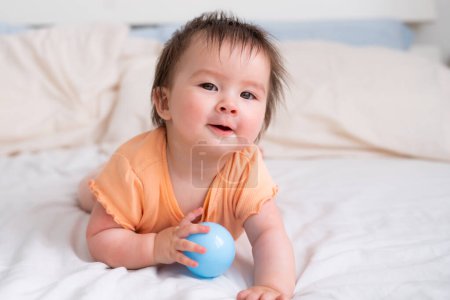 Téléchargez les photos : Mode de vie maison portrait de heureux et beau 8 mois bébé fille mixte race asiatique caucasien jouer avec ballon gai sur lit et explorer les environs curieux - en image libre de droit