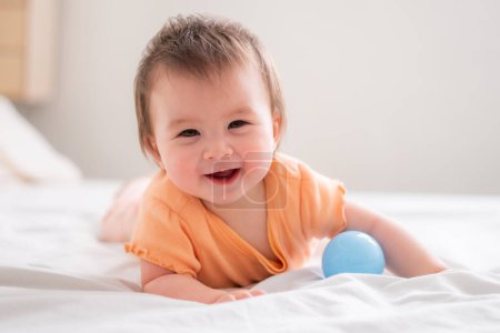 Téléchargez les photos : Mode de vie maison portrait de heureux et beau 8 mois bébé fille mixte race asiatique caucasien jouer avec ballon gai sur lit et explorer les environs curieux - en image libre de droit
