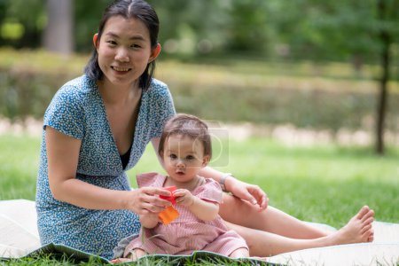 Foto de Estilo de vida al aire libre retrato de la madre y la hija - joven mujer china asiática feliz y dulce jugando con su bebé de 8 meses en la hierba en el parque de la ciudad - Imagen libre de derechos