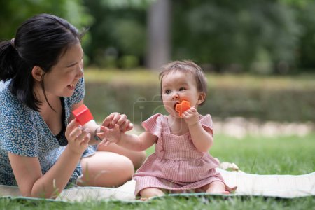 Foto de Estilo de vida al aire libre retrato de la madre y la hija - joven mujer china asiática feliz y dulce jugando con su bebé de 8 meses en la hierba en el parque de la ciudad - Imagen libre de derechos