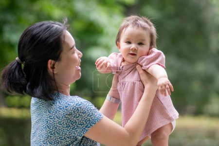 Téléchargez les photos : Portrait de mode de vie en plein air de la mère et de la fille - jeune femme coréenne asiatique heureuse et douce jouant avec son bébé fille de 8 mois au parc de la ville - en image libre de droit