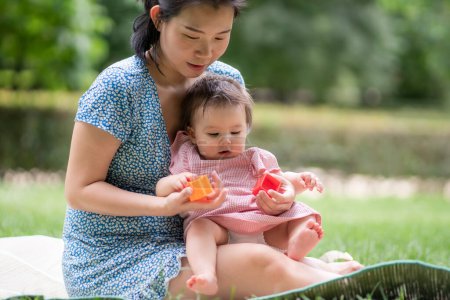 Foto de Estilo de vida al aire libre retrato de madre e hija - joven mujer coreana asiática feliz y dulce jugando con su bebé de 8 meses en el césped en el parque de la ciudad - Imagen libre de derechos