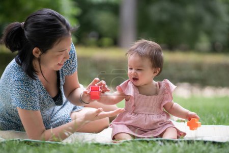 Téléchargez les photos : Portrait de mode de vie en plein air de la mère et de la fille - jeune femme coréenne asiatique heureuse et douce jouant avec son bébé fille de 8 mois sur l'herbe au parc de la ville - en image libre de droit