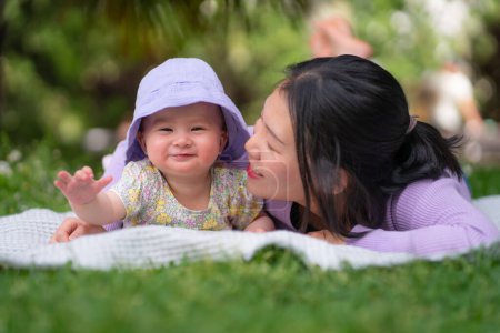 Foto de En el parque de la ciudad una niña de 5 meses feliz y adorable y su mujer china asiática comparten un momento lúdico en una manta en medio de la exuberante hierba verde en el amor y la conexión de la hija madre - Imagen libre de derechos