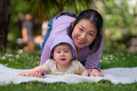 Foto de En el parque de la ciudad una niña de 5 meses feliz y adorable y su mujer coreana asiática comparten un momento lúdico en una manta en medio de la exuberante hierba verde en el amor y la conexión de la hija madre - Imagen libre de derechos