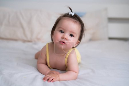 Téléchargez les photos : Ethnique mixte asiatique bébé fille caucasienne rampant sur le lit heureux et curieux - doux et adorable petit bébé de 8 mois avec une queue de cheval drôle explorant autour gai - en image libre de droit