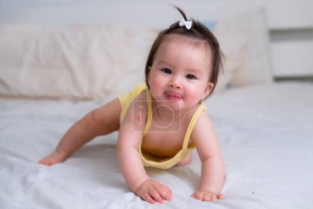 Téléchargez les photos : Ethnique mixte asiatique bébé fille caucasienne rampant sur le lit heureux et curieux - doux et adorable petit bébé de 8 mois avec une queue de cheval drôle explorant autour gai - en image libre de droit