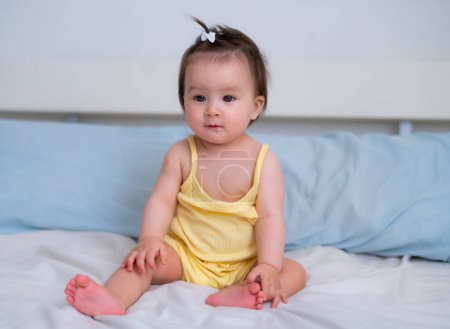 Téléchargez les photos : Ethnique mixte Asiatique Caucasien bébé fille assise sur le lit doux et adorable petit bébé 8 mois avec une queue de cheval drôle regardant autour curieux - en image libre de droit