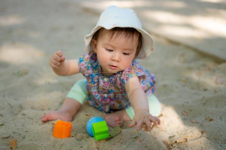 Foto de Adorable bebé de 9 meses jugando al aire libre - retrato de estilo de vida de etnia mixta niña asiática caucásica jugando con juguetes de bloque feliz y despreocupado en el patio de recreo sentado en la arena - Imagen libre de derechos