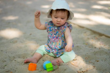Foto de Adorable bebé de 9 meses jugando al aire libre - retrato de estilo de vida de etnia mixta niña asiática caucásica jugando con juguetes de bloque feliz y despreocupado en el patio de recreo sentado en la arena - Imagen libre de derechos