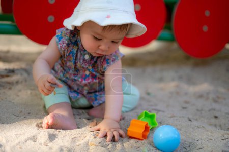 Photo for Adorable 9 months old baby playing outdoors - lifestyle portrait of mixed ethnicity Asian Caucasian baby girl playing with block toys happy and carefree at playground sitting on sand - Royalty Free Image