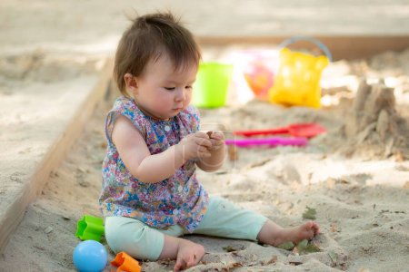 Téléchargez les photos : Adorable bébé de 9 mois jouant à l'extérieur portrait style de vie d'origine ethnique mixte asiatique Caucasien bébé fille jouant avec des jouets bloc heureux et insouciant à aire de jeux assis sur le sable - en image libre de droit