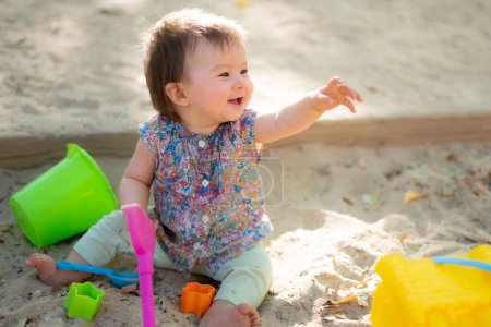 Photo for Adorable 9 months old baby playing outdoors - lifestyle portrait of mixed ethnicity Asian Caucasian baby girl playing with block toys happy and carefree at playground sitting on sand - Royalty Free Image