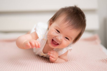 Téléchargez les photos : Mode de vie portrait à la maison de 9 mois mélange ethnique asiatique Caucasien bébé fille jouer heureux et insouciant sur lit rampant rire et excité dans l'enfance et la crèche concept - en image libre de droit