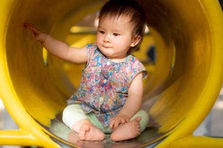 Téléchargez les photos : Portrait de style de vie en plein air de belle et adorable bébé fille de 9 mois assis sur tube jaune à aire de jeux pour enfants dans le parc de la ville explorer les environs joyeux et curieux - en image libre de droit