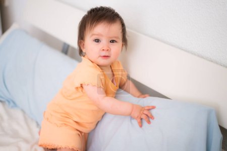 Téléchargez les photos : Mode de vie portrait à la maison de heureuse et adorable fille de 9 mois regardant la caméra dans une expression drôle de visage sur fond blanc rampant sur le lit - en image libre de droit