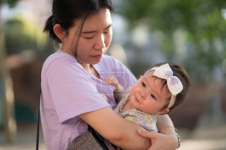 Téléchargez les photos : Portrait de style de vie en plein air de la femme chinoise asiatique tenant belle et adorable bébé fille souriant gai sur aire de jeux portant un ruban mignon dans le concept de liaison mère fille - en image libre de droit