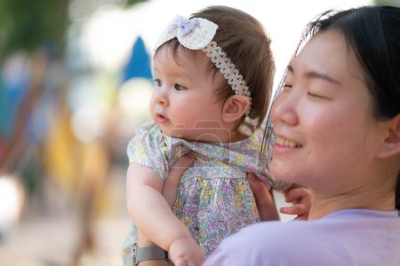 Téléchargez les photos : Portrait de style de vie en plein air de la femme chinoise asiatique tenant belle et adorable bébé fille souriant gai sur aire de jeux portant un ruban mignon dans le concept de liaison mère fille - en image libre de droit
