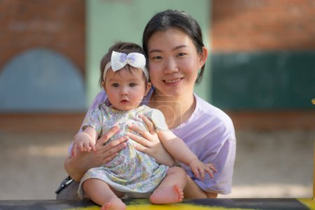 Photo for Outdoors lifestyle portrait of Asian Chinese woman playing with beautiful and adorable baby girl smiling cheerful on playground playing on swing at city park wearing a cute ribbon - Royalty Free Image