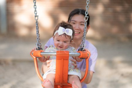 Téléchargez les photos : Portrait de style de vie en plein air de femme asiatique jouant avec belle et adorable petite fille souriant gai sur aire de jeux jouant sur swing au parc de la ville portant un ruban mignon dans le concept de l'enfance - en image libre de droit