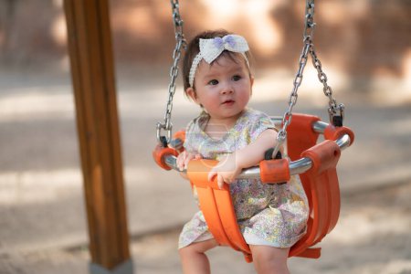 Téléchargez les photos : Portrait de style de vie en plein air de belle et adorable 8 ou 9 mois bébé fille souriant gai sur aire de jeux jouant sur swing au parc de la ville portant un ruban mignon dans le concept de l'enfance - en image libre de droit