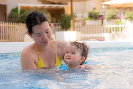 Téléchargez les photos : Heureuse et belle femme asiatique tenant sa petite fille ludique mère coréenne et adorable fille jouant sur l'eau à la piscine de villégiature en été - en image libre de droit