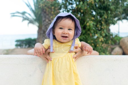 Photo for Outdoors lifestyle portrait of adorable and happy baby girl in cute hat hold by her mothers hands on park bench looking excited and cheerful during Summer holidays - Royalty Free Image
