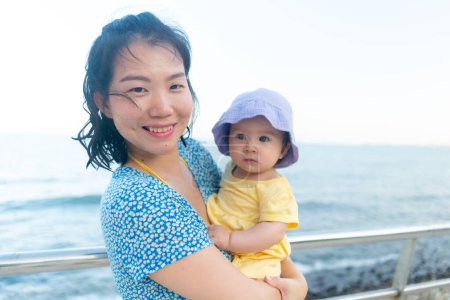 Téléchargez les photos : Portrait de mode de vie en plein air de la mère asiatique et sa petite fille belle femme tenant son adorable et heureuse petite fille à la mer profitant des vacances d'été - en image libre de droit