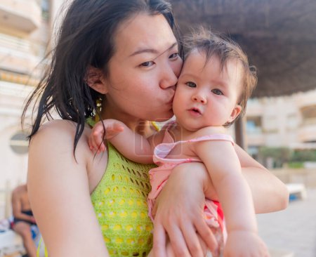 Foto de Mujer asiática besando a su adorable bebé - hermosa madre cariñosa coreana abrazando a su hija pequeña abrazándola fuerte y besándola durante las vacaciones de verano - Imagen libre de derechos