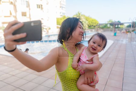 Foto de Mujer asiática tomando foto selfie con su adorable bebé - hermosa madre coreana sosteniendo a su hija pequeña y tomando una foto con el teléfono móvil durante las vacaciones - Imagen libre de derechos
