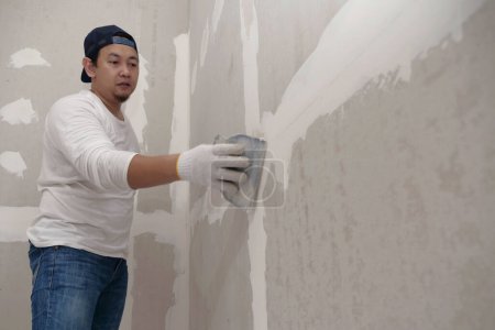 Photo for Plasterwork and wall painting preparation. Asian male applying plaster or filling drywall patch - Royalty Free Image