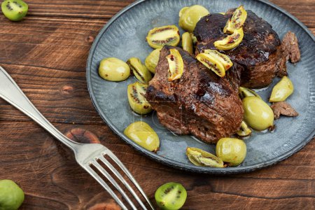 Téléchargez les photos : Plat de steak d'autruche rôti avec mini sauce kiwi. Viande aux baies de kiwi sur table en bois. - en image libre de droit