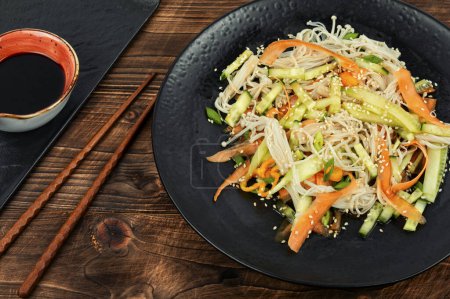 Téléchargez les photos : Salade saine avec champignons enoki, carottes, concombre et graines de sésame sur une table rustique en bois. Nourriture, salade servie sur une assiette noire. - en image libre de droit