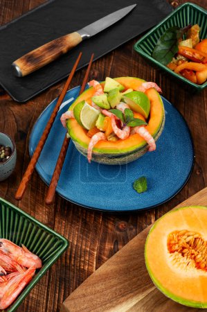 Salat, Obstsalat aus Garnelen, Melone und Avocado in Melone auf rustikalem Holztisch.