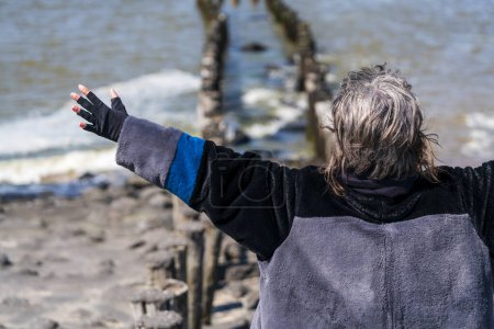 Photo for Mature woman stands with arms spread, gazing at the sea from behind standing to the right, braving the cold day. - Royalty Free Image