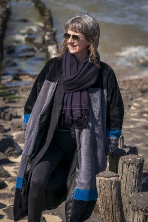 Eine reife Frau in Winterkleidung steht da und lächelt, während sie an der holländischen Küste mit dem Meer im Rücken dem kalten Tag trotzt.