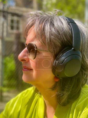 Une femme mature et belle à la hanche est assise dans un jardin printanier, vêtue d'une veste moderne verte et de lunettes de soleil, respirant la confiance et le style au milieu de la nature florissante.