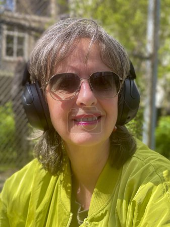 Portrait de femme mature et branchée assise dans un jardin printanier, vêtue d'une veste moderne verte et de lunettes de soleil, respirant la confiance et le style au milieu de la nature florissante.