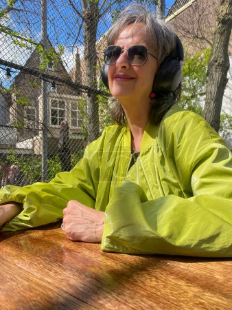 Eine reife, schöne hippe Frau sitzt in einem frühlingshaften Garten, trägt eine grüne, moderne Jacke und Sonnenbrille und strahlt inmitten der blühenden Natur Selbstvertrauen und Stil aus..