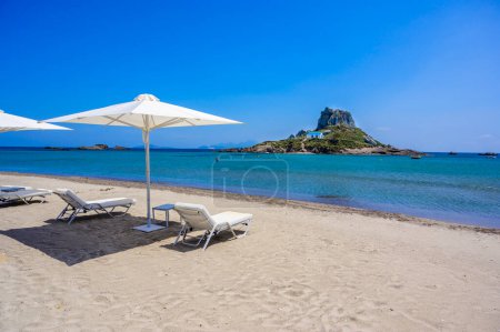 Téléchargez les photos : Chaise longue et parasol sur la belle plage d'Agios Stefanos en face du paradis Île de Kastri- ruines historiques et paysages paradisiaques à la côte de l'île de Kos, Grèce - en image libre de droit
